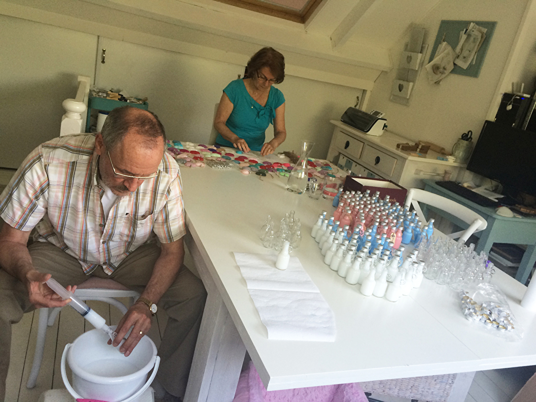 Twee mensen werken samen aan een tafel vol flesjes, zeepjes en creatieve materialen in een knutselruimte – met de hand vullen en voorbereiden van bedankjes of cadeautjes in huiselijke sfeer.