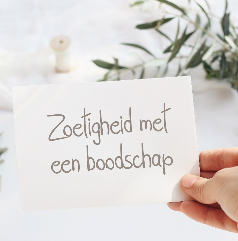 Wit kaartje met handgeschreven tekst "Zoetigheid met een boodschap", vastgehouden door een hand met op de achtergrond een zachte, natuurlijke styling