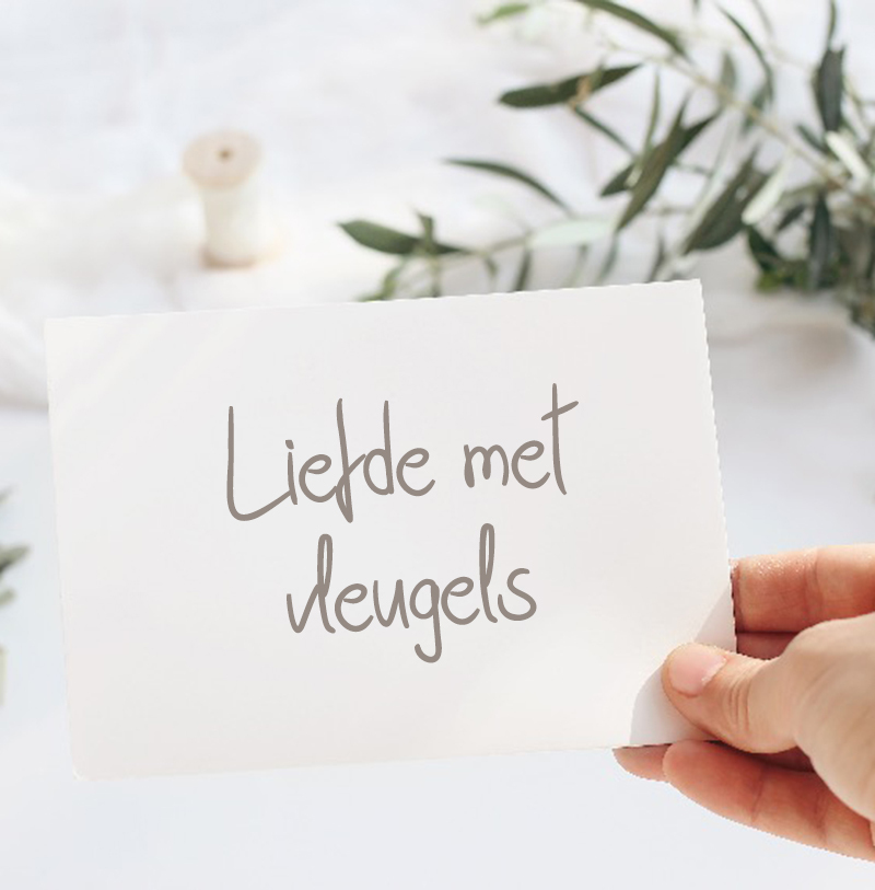 Hand met kaart waarop de tekst ‘Liefde met vleugels’ staat, tegen een zachte achtergrond met olijftakken en garen
