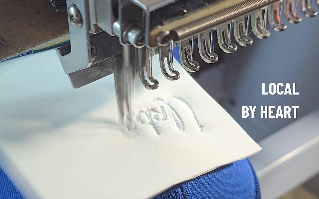Close-up van een industri&euml;le borduurmachine die het witte Urbs Roffa logo in 3D borduurt op een blauwe cap.