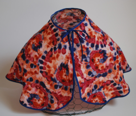 Een kapmanteltje van crêpe met een fantasie bloemmotief in de kleuren rood, oranje en blauw, met blauw biasband. Nog 1 op voorraad.