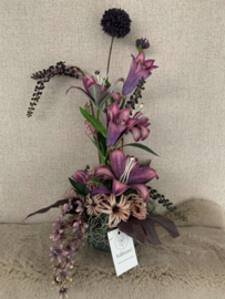 Bloemstuk in ruwe grijze pot met mauve lelie's, paarse bessen, roze clematis, mauve wisteria met goud, bruin/paars blad, grijze tillandsia